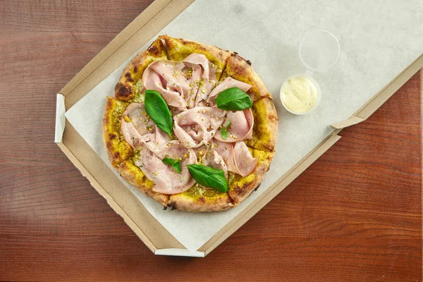 Photo for pizza delivery. Pizza with mortadella sausage, pistachios, basil and melted cheese in a cardboard box with side sauce on a wooden table