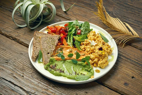 An appetizing vegetarian dish is scramble from tofu cheese, a salad of baked peppers and arugula and two types of hummus. Wood background