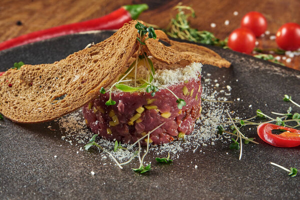 Close up view on fresh beef tartare with bread, egg and microgreen in black plate on wooden background.