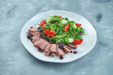 Domatesli fırında ördek göğsü salatası, roka, gri arka planda beyaz bir tabakta yapraklar.