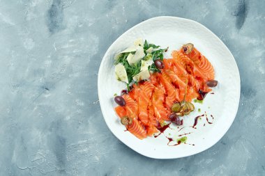 Hafif tuzlu somon dilimlenmiş roka ve parmesanlı. Kırmızı balıklı sashimi kapari ve gri arka planda beyaz bir tabakta zeytin taze deniz ürünleri.