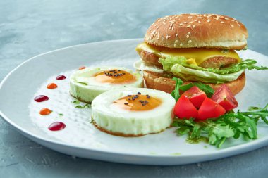 Mükemmel kahvaltı - tavuk ve peynir burger, 2 sahanda yumurta ve roka ve kiraz domatesli salata gri arka planda beyaz bir tabakta