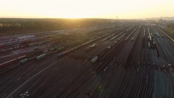 Latający widok z powietrza na kolejową stację sortującą o zachodzie słońca. Wiele wagonów i torów kolejowych na przemysłowej stacji towarowej. Pociąg porusza się wzdłuż torów. Piękny zachód słońca. Przemysł ciężki — Wideo stockowe