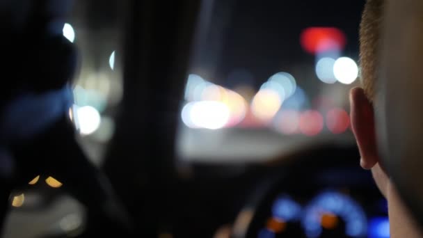 Un conductor que conduce un coche contra el fondo de una carretera nocturna y el resplandor de los coches que pasan. Primer plano. Vista desde el asiento trasero. Hermoso bokeh colorido. 4K en cámara lenta — Vídeos de Stock