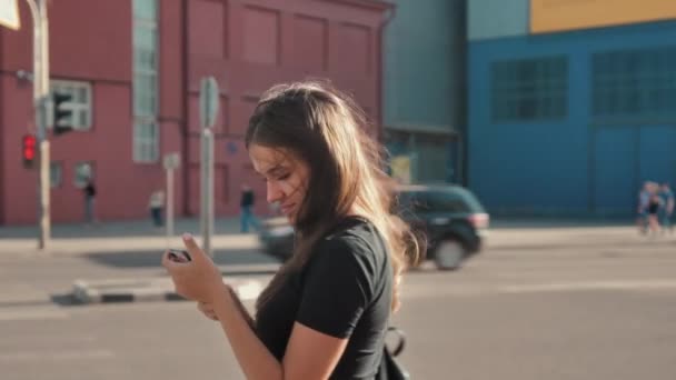 Uzun saçlı güzel esmer, yaya geçidinin önünde akıllı telefon kullanıyor. Çekici genç kadınlar güneşli bir şehrin sokaklarında yürüyor ve gülümsüyor ve arkadaşlarıyla sohbet ediyorlar. — Stok video