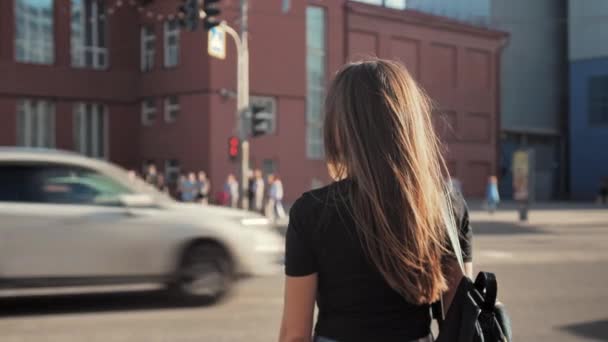 Widok z tyłu. Młoda brunetka czeka na zielone światło, aby przejść przez ulicę w mieście. Samochody jadą wzdłuż drogi. Letni słoneczny dzień. — Wideo stockowe