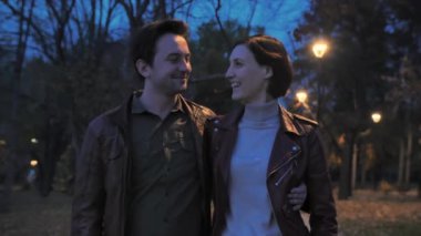 Akşam parkında yürürken sevimli mutlu çift kucaklaşması. Karantinadan sonra yürüyün. Eğlenin, gülün, sohbet edin, gün batımını izleyin. Gençler şehrin arka planında yürüyorlar. Yavaş