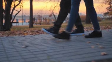 Genç bir çift sonbahar parkında güzel bir günbatımının arka planında yürüyor. Sur boyunca yürüyen iki romantik insanın bacakları dinlenen insanların arka planına karşı. Mutlu
