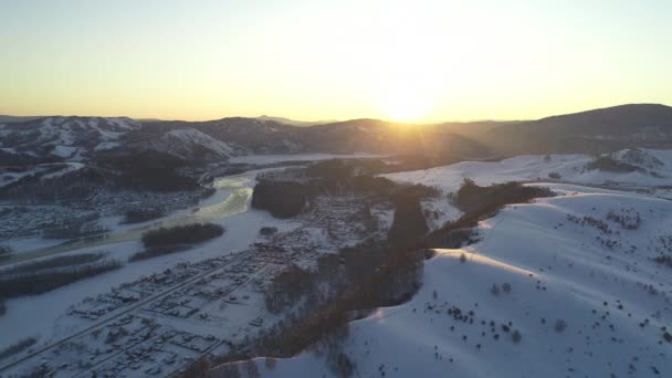 Vista aérea: Voo sobre a aldeia junto ao rio no inverno. Belo nascer do sol nas montanhas. Telhados de casas cobertos de neve. Tempo gelado. Paisagem de inverno, natureza, raios de sol. Temporada de viagem. Neve. — Vídeo de Stock