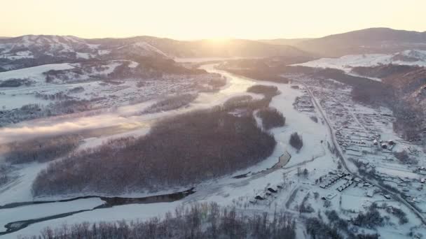 Voo sobre o rio de inverno nas montanhas. Belo nascer do sol por detrás das montanhas. Névoa sobre o rio. Floresta nevada e uma aldeia junto ao rio. Paisagem de inverno, natureza, raios de sol. Viagens — Vídeo de Stock