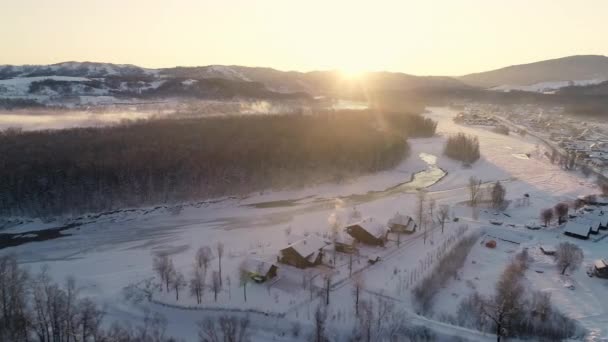 Uitzicht vanuit de lucht: vlucht langs een bevroren rivier in de bergen. Prachtige zonsopgang. Een dorp aan de kust, met sneeuw bedekte daken van huizen, rook uit een schoorsteen. Winterlandschap, natuur, zonnestralen. Reizen — Stockvideo