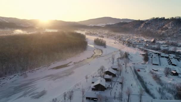 Vista aérea: voo sobre a aldeia junto ao rio no inverno. Belo nascer do sol nas montanhas. Telhados de casas cobertos de neve. Gelo no rio. Paisagem de inverno, natureza, raios de sol. Temporada de viagem — Vídeo de Stock