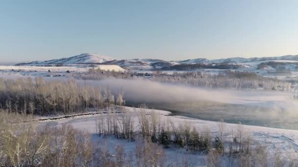 空中ビュー:冬の川の上に朝の霧。川に氷が浮かんでいる。背景に美しい山々、霜の木。海岸沿いの村。冬の風景、自然、太陽光線。旅行 — ストック動画