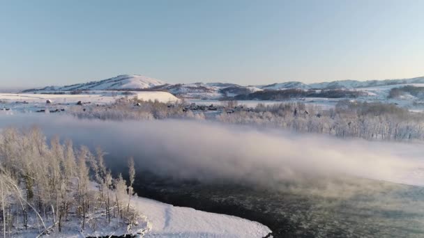 Vlucht over de winterochtendmist over de rivier. IJs drijft op de rivier. Prachtige bergen op de achtergrond, bomen in hevige vorst. Dorp aan de kust. Winterlandschap, natuur, zonnestralen. Reizen — Stockvideo