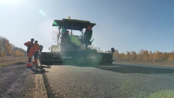 Novosibirsk, 3 de agosto de 2020. Construcción de una nueva carretera. Reparación de carreteras. Los trabajadores con overoles están trabajando con una pavimentadora de asfalto. Una capa de asfalto recién puesto al atardecer. Vapor de caliente — Vídeo de stock