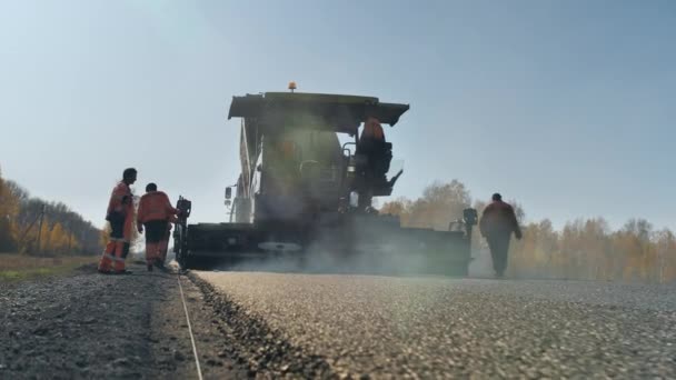 Novosibirsk, 3 de agosto de 2020. Construcción de una nueva carretera. Los trabajadores con overoles están trabajando con una pavimentadora de asfalto. Una capa de asfalto recién puesto al atardecer. Asfalto caliente. Hermoso resplandor. — Vídeos de Stock
