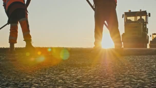 Two road workers in overalls level the fresh asphalt mix against the background of road rollers. Silhouettes of men in the rays of the setting sun. Road repair, hot asphalt, bitumen. — Stock Video