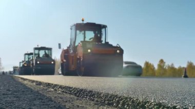 Yeni yerleştirilmiş asfalt tabakası. Yol yüzeyi tamiri. Yeni bir yol inşaatı. Patenler mavi gökyüzüne karşı asfalt seviyesini düşürür ve sıkıştırır. Yol silindirleri. Güneşli bir gün.