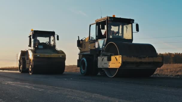 Riparazione della superficie stradale. Costruzione di una nuova strada. Tre rulli livellano e compattano l'asfalto al tramonto, le auto passano. Uno strato di asfalto appena deposto nei raggi del sole al tramonto. — Video Stock