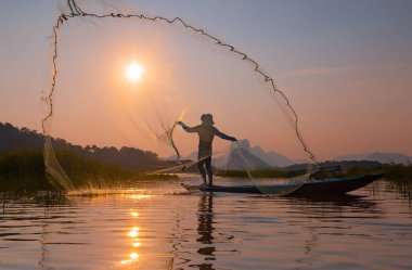 Kimliği belirsiz balıkçı silueti gün batımında alacakaranlık gökyüzü ile barajdaki ahşap tekneye ağ atıyor. 
