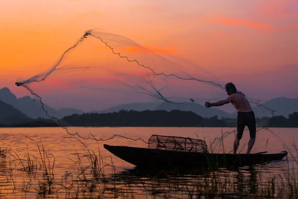 Tanımlanamayan bir balıkçı, gün batımında alacakaranlık gökyüzü ile birlikte barajdaki ahşap tekneye ağ atıyor..