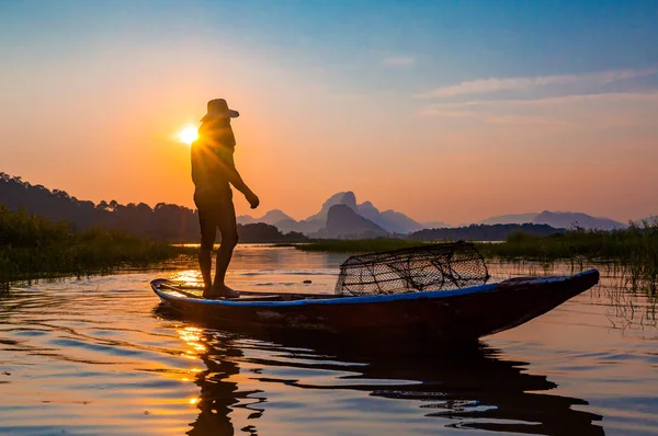 Kimliği belirsiz balıkçı silueti gün batımında alacakaranlık gökyüzü ile barajdaki ahşap tekneye ağ atıyor. 