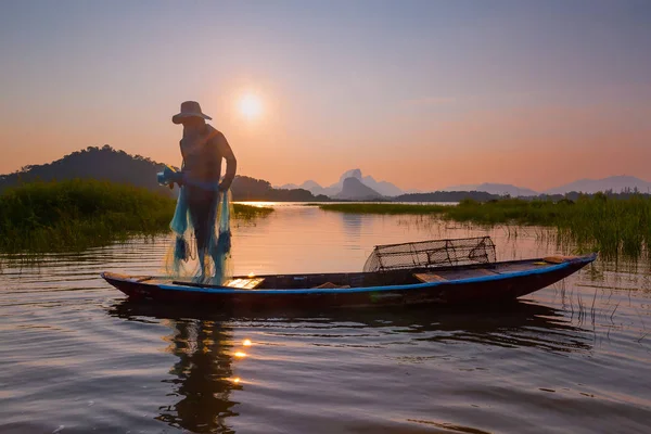 Kimliği belirsiz balıkçı silueti gün batımında alacakaranlık gökyüzü ile barajdaki ahşap tekneye ağ atıyor. 