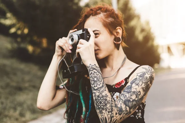 young girl with tattoos and dreadlocks photographs vintage camera in the park
