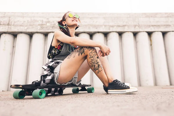 Young girl with tattoo and dreadlocks sitting on a longboard on the background of concrete structure — Stock Photo, Image
