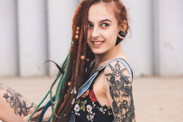 young girl with tattoo and dreadlocks is sitting on the background of concrete structure