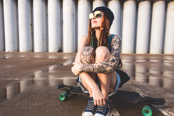 Young girl with tattoos and dreadlocks in a blue cap sits on a longboard against the background of concrete structure — Stock Photo, Image