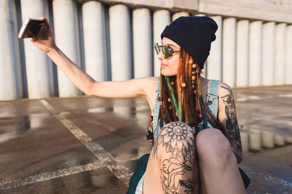 Chica joven con tatuajes y rastas en una gorra azul utiliza un teléfono inteligente contra el fondo de una pared de hormigón —  Fotos de Stock