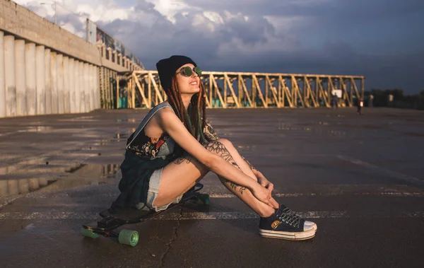 Jovem com tatuagens e dreadlocks em um boné azul senta-se em um longboard contra o fundo da estrutura de concreto — Fotografia de Stock