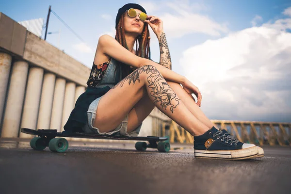 Chica joven con tatuajes y rastas en una gorra azul se sienta en un longboard sobre el fondo de la estructura de hormigón — Foto de Stock