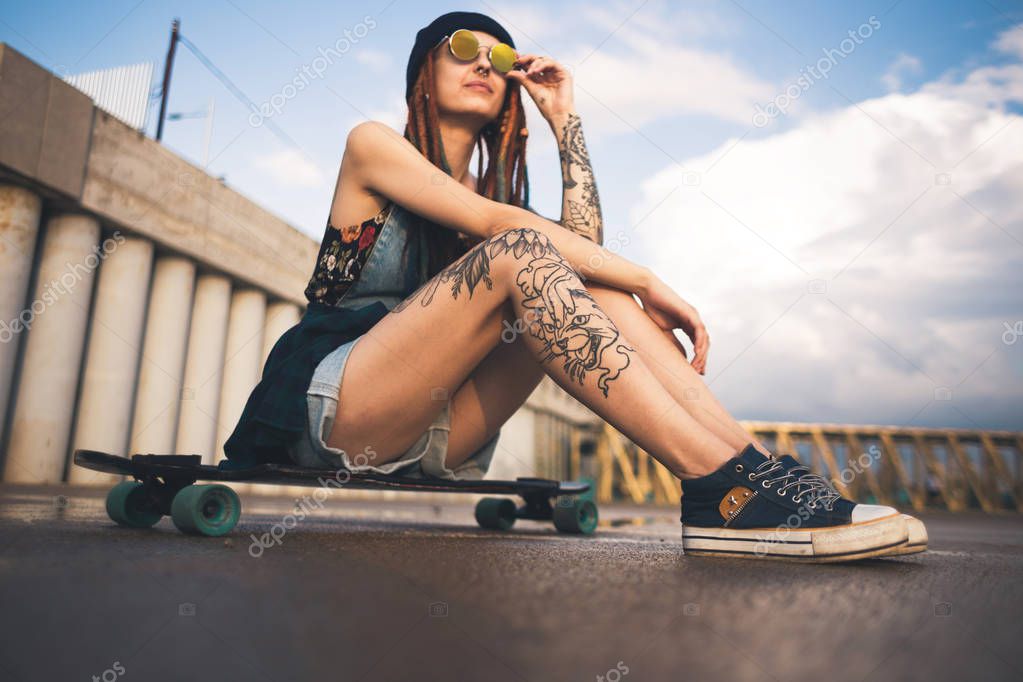 young girl with tattoos and dreadlocks in a blue cap sits on a longboard against the background of concrete structure