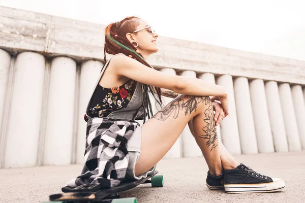 Jovem com tatuagem e dreadlocks sentado em um longboard no fundo da estrutura de concreto — Fotografia de Stock