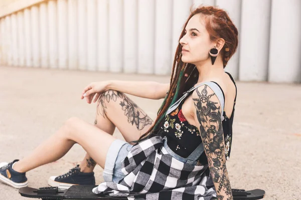 Молодая девушка с татуировкой и дредами, сидящая на доске на фоне бетонной конструкции — стоковое фото