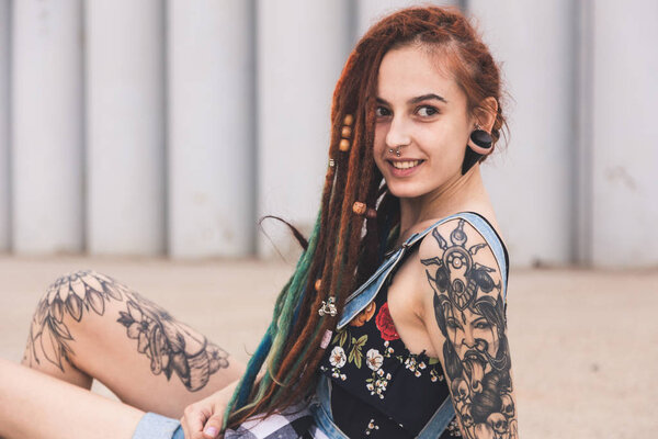 young girl with tattoo and dreadlocks is sitting on the background of concrete structure