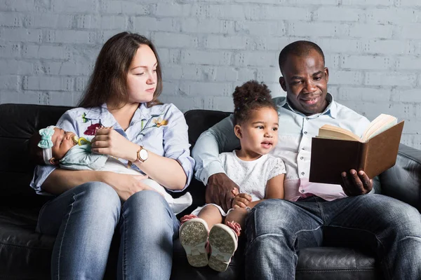 Parents read a book to children sitting on the couch. Happy multiethnic family. Family values