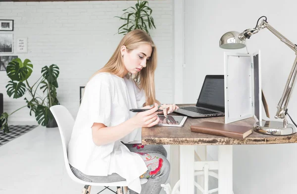 Νεαρή γυναίκα που εργάζονται στο σπίτι πίσω από ένα φορητό υπολογιστή και tablet. Δημιουργική σκανδιναβικό στυλ χώρου εργασίας — Φωτογραφία Αρχείου