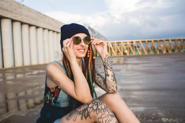 Chica joven con tatuajes y rastas en una gorra azul se sienta en el fondo de la estructura de hormigón —  Fotos de Stock