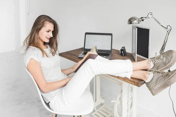Νεαρή γυναίκα εργασία στο σπίτι και διαβάζοντας ένα βιβλίο με τα πόδια της πάνω στο τραπέζι. Δημιουργική σκανδιναβικό στυλ χώρου εργασίας. — Φωτογραφία Αρχείου