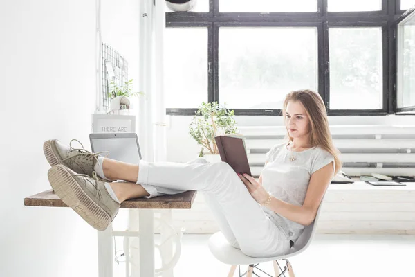 Joven mujer de negocios trabajando en casa y leyendo un libro con las piernas sobre la mesa. Espacio de trabajo de estilo escandinavo creativo — Foto de Stock