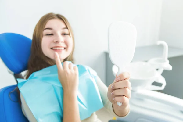 Clínica dental. Recepción, examen del paciente. Cuidado de los dientes. Chica joven sonriendo, mirando en el espejo después de un chequeo dental a su dentista —  Fotos de Stock