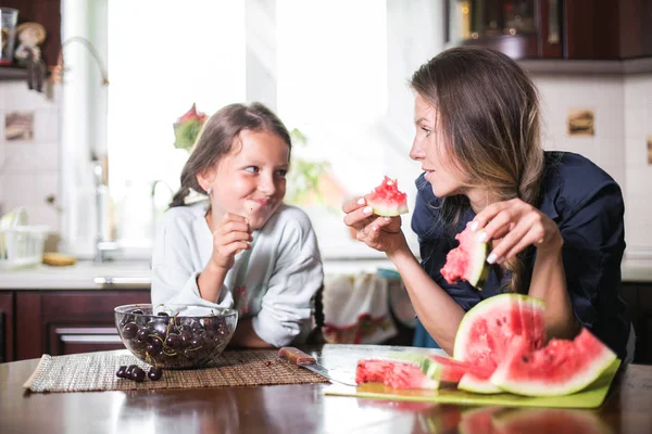 Linda niña y su hermosa mamá están cortando frutas, sandía roja y sonriendo mientras cocinan en la cocina en casa. Familia feliz — Foto de Stock