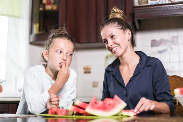 Linda niña y su hermosa mamá están cortando frutas, sandía roja y sonriendo mientras cocinan en la cocina en casa. Familia feliz — Foto de Stock