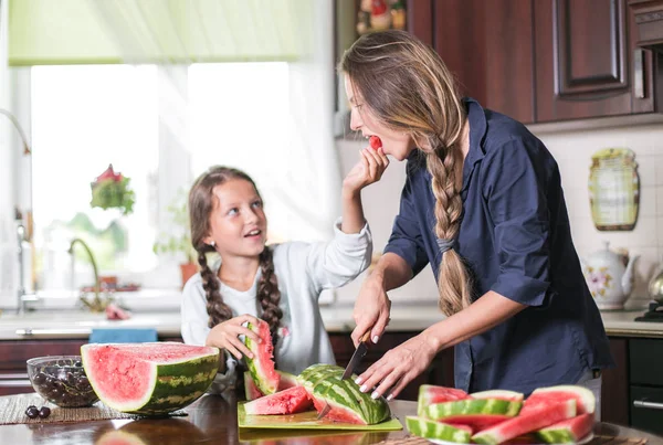 Linda niña y su hermosa mamá están cortando frutas, sandía roja y sonriendo mientras cocinan en la cocina en casa. Familia feliz — Foto de Stock