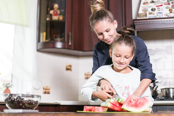 Linda niña y su hermosa mamá están cortando frutas, sandía roja y sonriendo mientras cocinan en la cocina en casa. Familia feliz — Foto de Stock