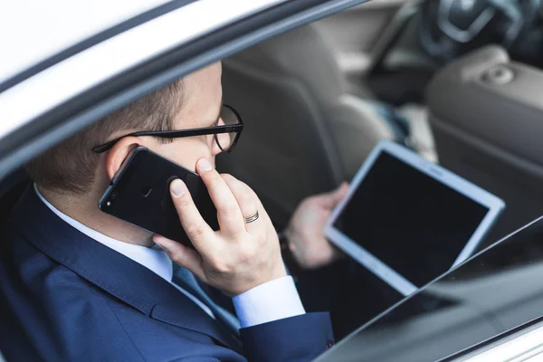 Młody biznesmen rozmowy na telefon komórkowy i przy użyciu komputera typu tablet siedząc na tylnym siedzeniu samochodu. Kaukaski mężczyzna firmy wykonawczej przez taksówkę i patrząc cyfrowych tabletu. — Zdjęcie stockowe