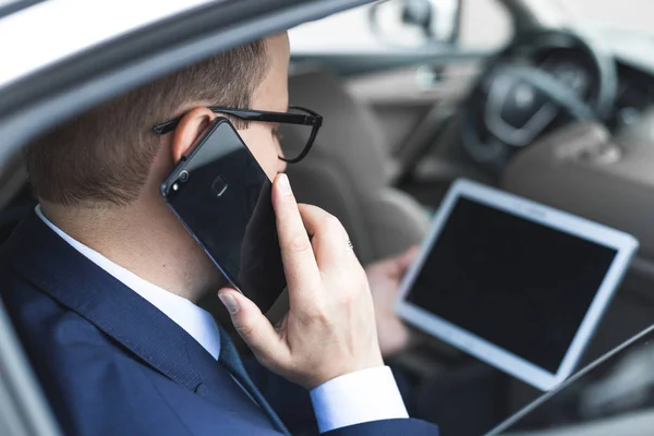Młody biznesmen rozmowy na telefon komórkowy i przy użyciu komputera typu tablet siedząc na tylnym siedzeniu samochodu. Kaukaski mężczyzna firmy wykonawczej przez taksówkę i patrząc cyfrowych tabletu. — Zdjęcie stockowe